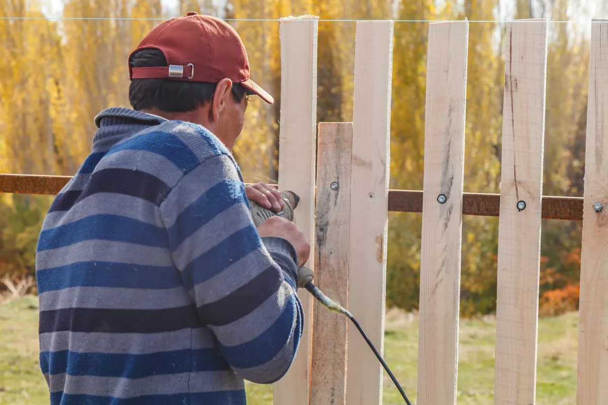 A man uses a drill to tighten a screw while installing a picket fence, showcasing fence maintenance.