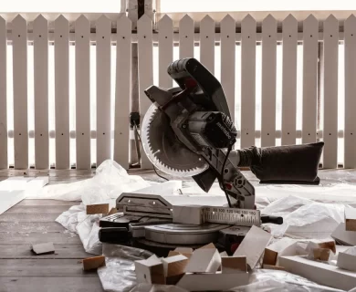 A circular saw on a wooden terrace with cut wood pieces and a white fence, suggesting DIY fence repair.