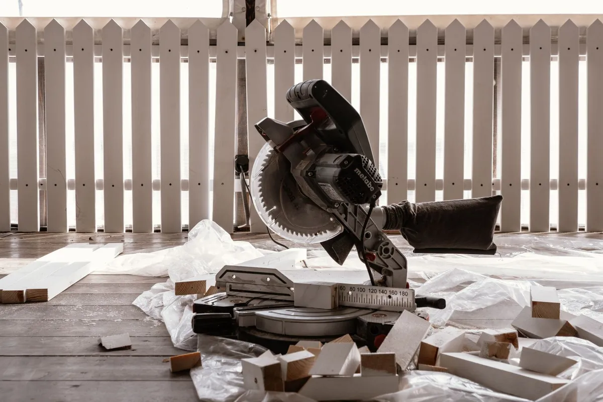 A circular saw on a wooden terrace with cut wood pieces and a white fence, suggesting DIY fence repair.